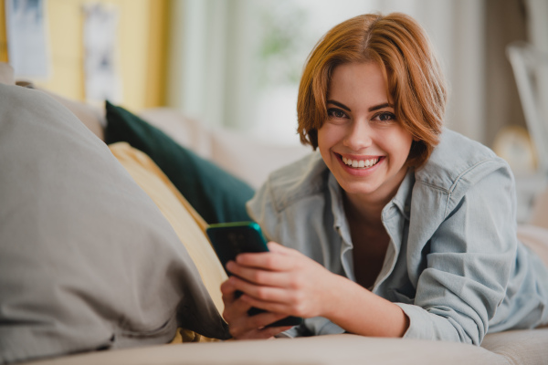 Portrait of a happy young woman using smartphone, lying on sofa at home, looking at camera, social networks concept.