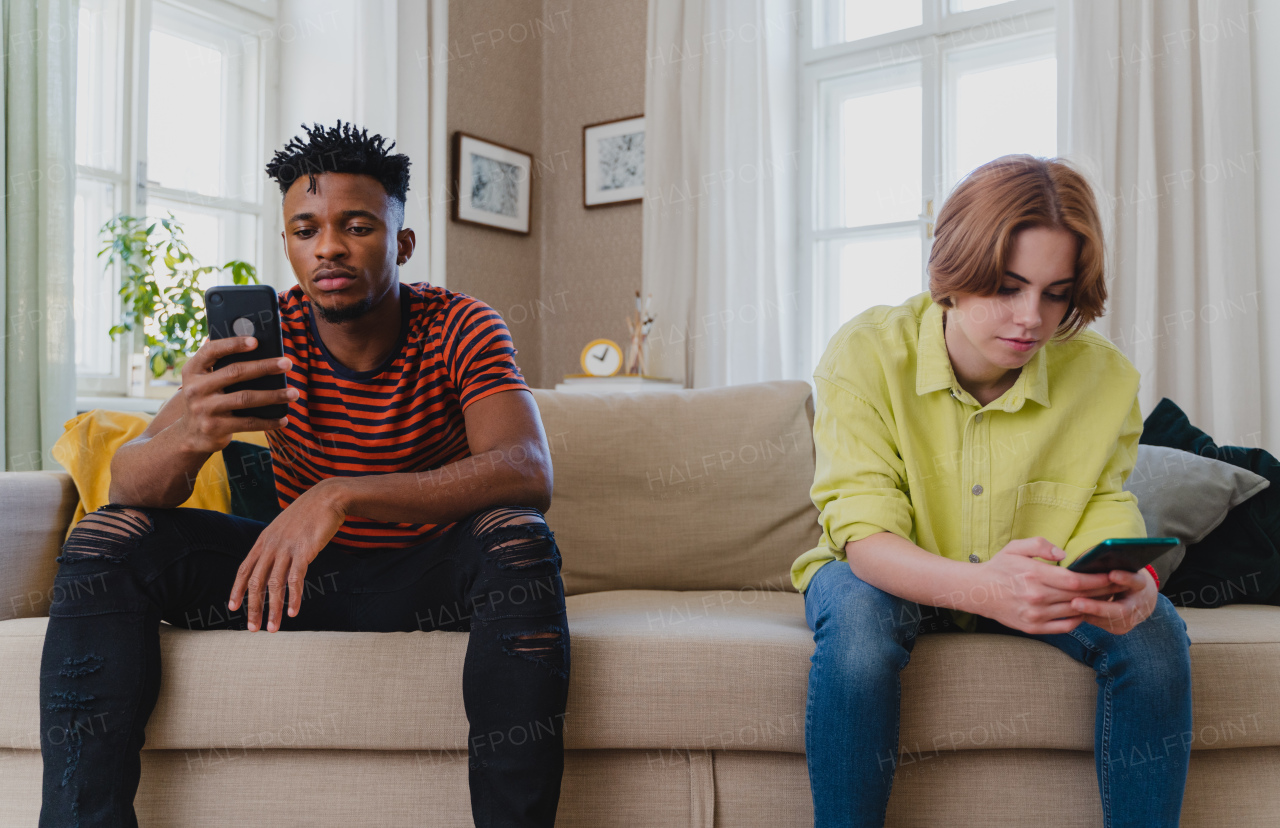 Young biracial couple addicted to smartphones sitting on a sofa at home, social media concept.