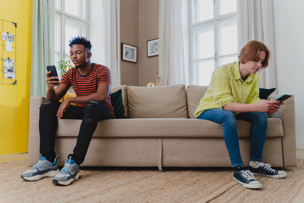 Young mixed couple addicted to smartphones ignoring each other sitting on sofa at home. Relationship problems