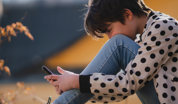 A side view of preteen girl addicted to smartphone sitting outdoors in nature