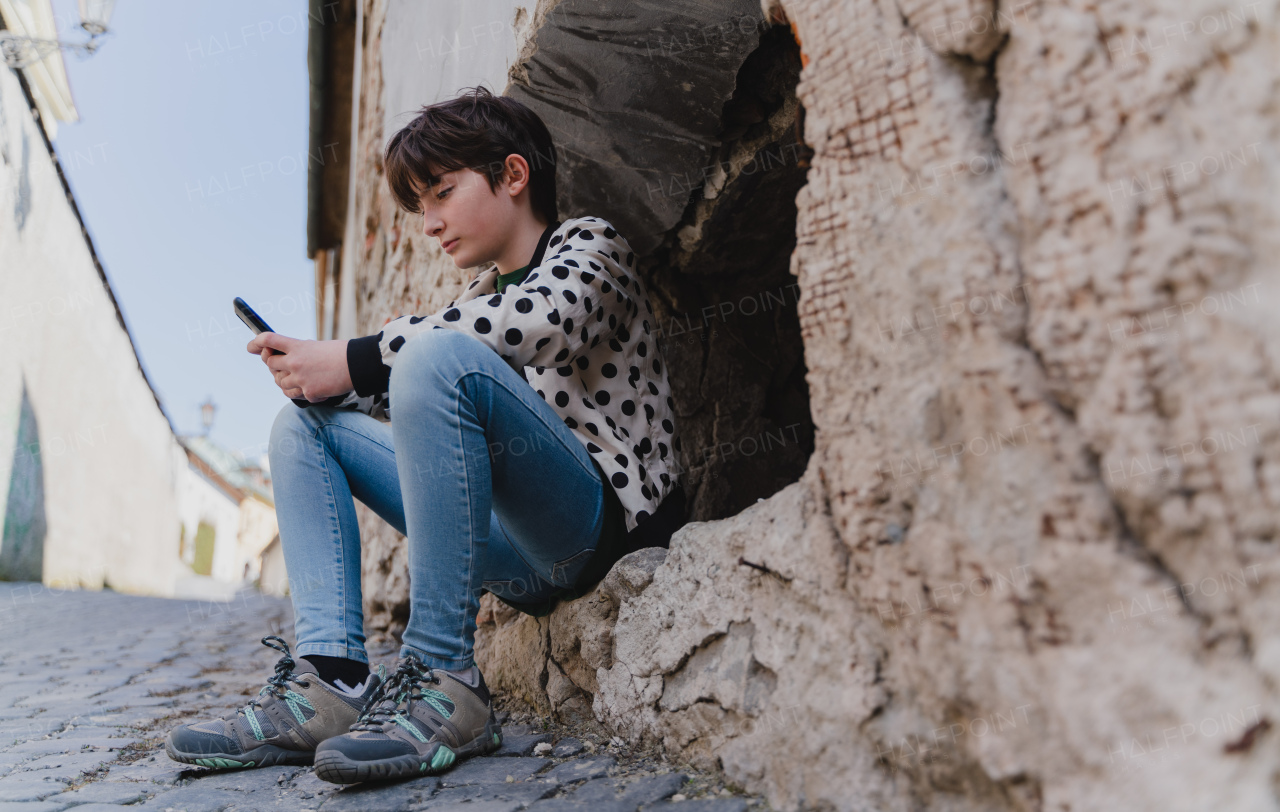 A low angle view of preteen girl with smartphone sitting outdoors in town.