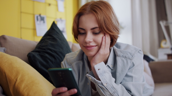 A cheerful teenager laying on sofa, using smartphone and looking at camera. Social networks concept.