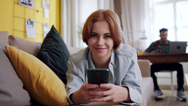 A cheerful teenager laying on sofa, using smartphone and looking at camera. Social networks concept.