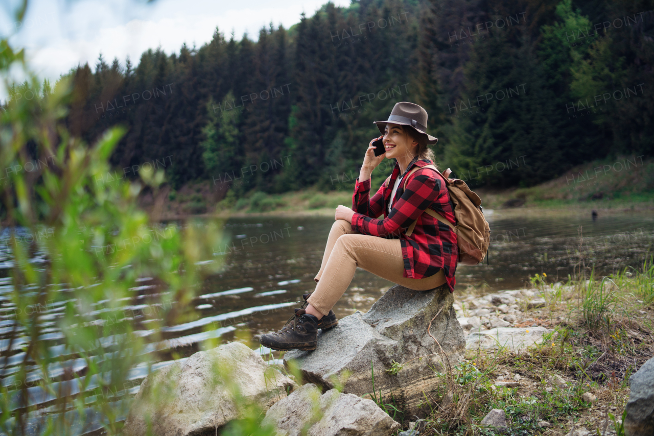 Side view of young woman sitting by lake on a walk outdoors in summer nature, using telephone.