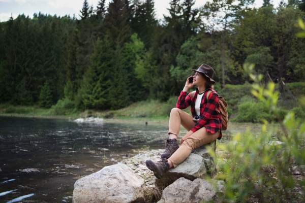 Side view of young woman sitting by lake on a walk outdoors in summer nature, using telephone.