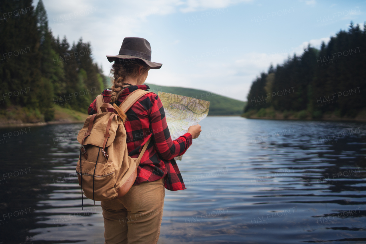 A rear view of young woman standing by lake outdoors in summer nature, using map.