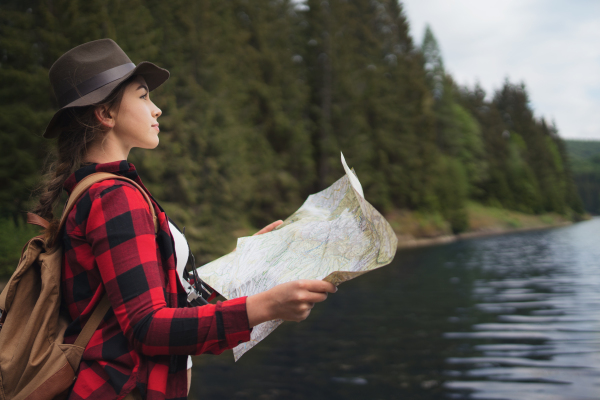 Young woman with closed eyes standing by lake on a walk outdoors in summer nature, using map.