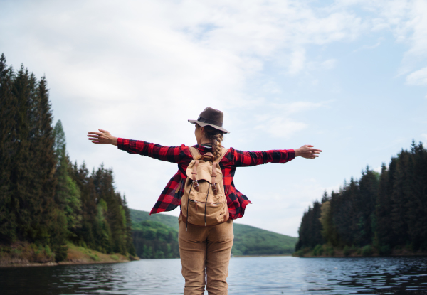 Rear view of young woman standing by lake on a walk outdoors in summer nature, relaxing.