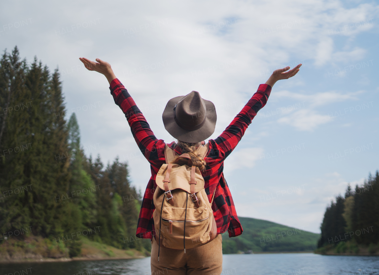 Rear view of young woman standing by lake on a walk outdoors in summer nature, relaxing.