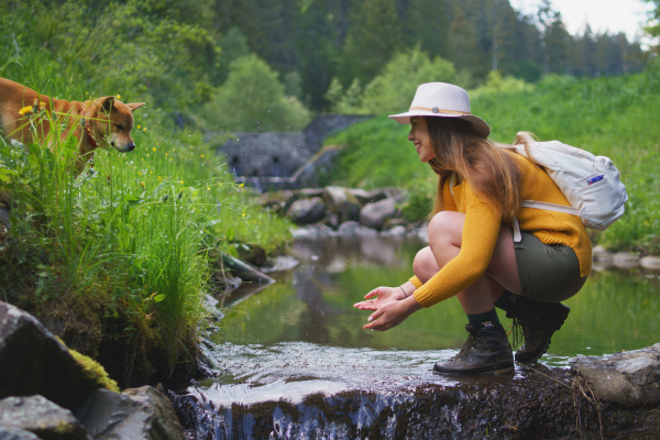 Happy young woman with dog standing by stream on a walk outdoors in summer nature, washing hands.