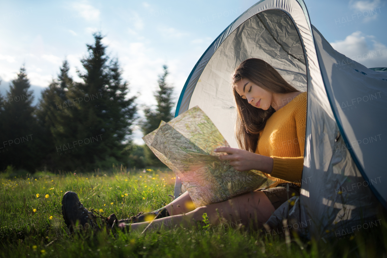 Side view of happy young woman sitting in tent shelter outdoors in summer nature, using map.