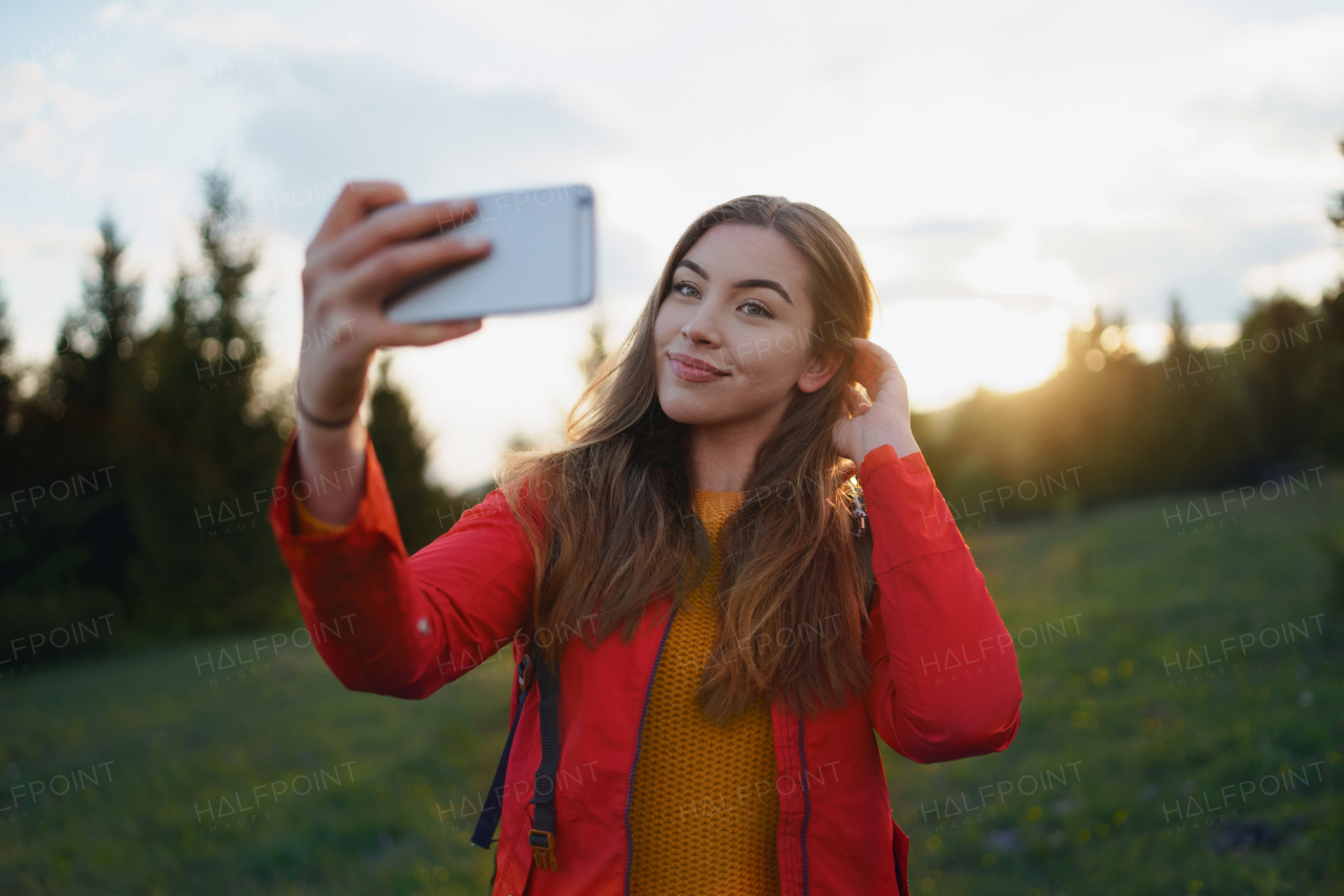 Side view of young woman on a walk outdoors on meadow in summer nature, taking selfie.