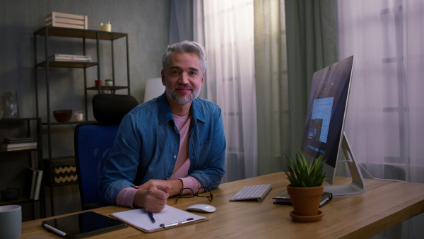 A mature businessman working on laptop at desk indoors in office, looking at camera.