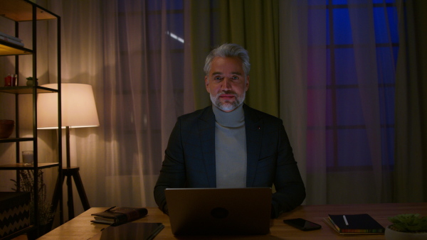 A mature businessman working on laptop at desk indoors in office, looking at camera.