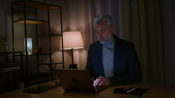 A mature businessman working on laptop at desk indoors in office, looking at camera.