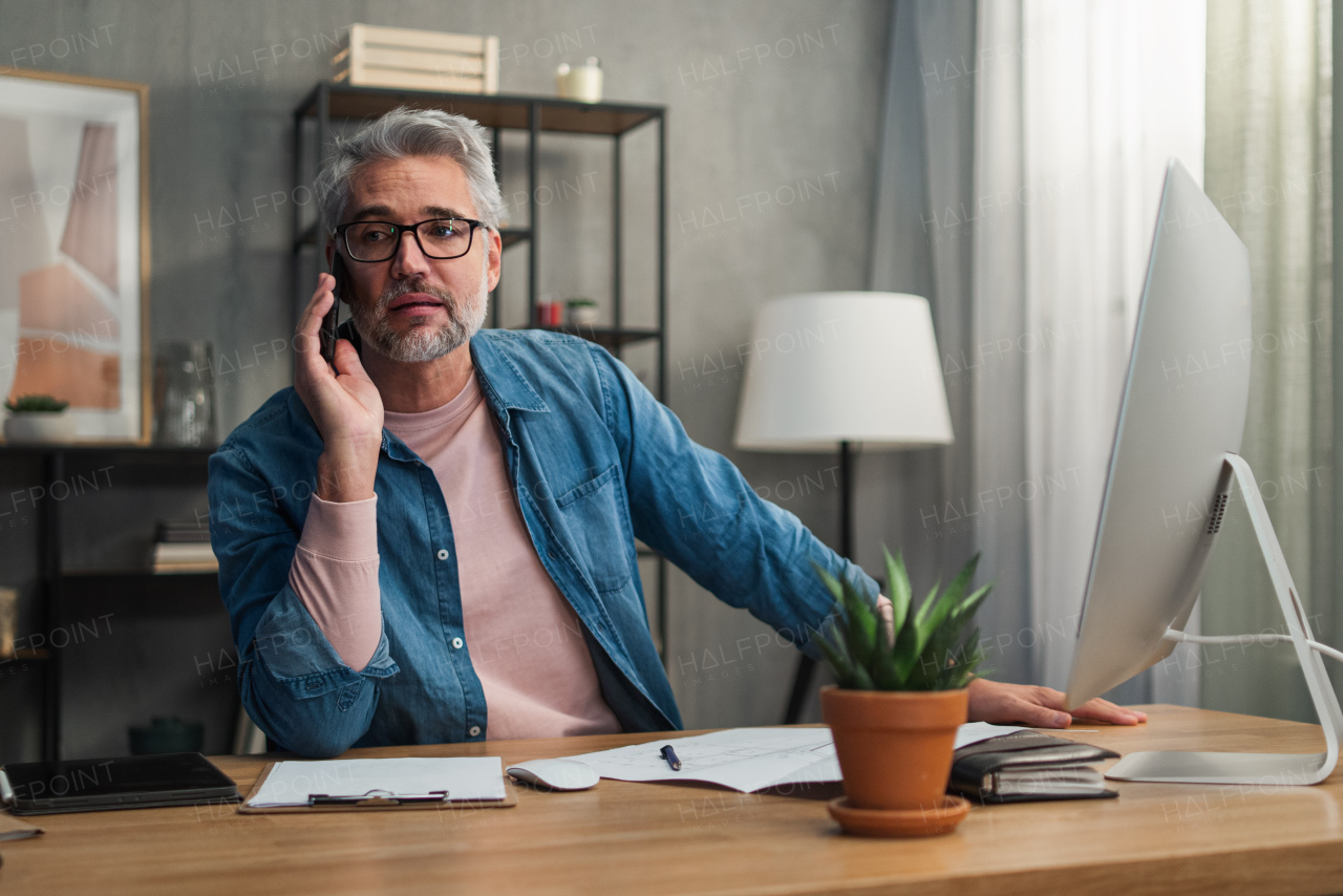 A mature man architect working on computer and making phone call at desk indoors in office.