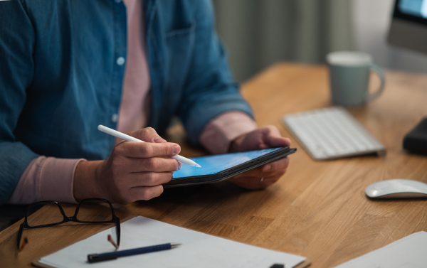 A close-up of mature man working on tablet at desk indoors in office.