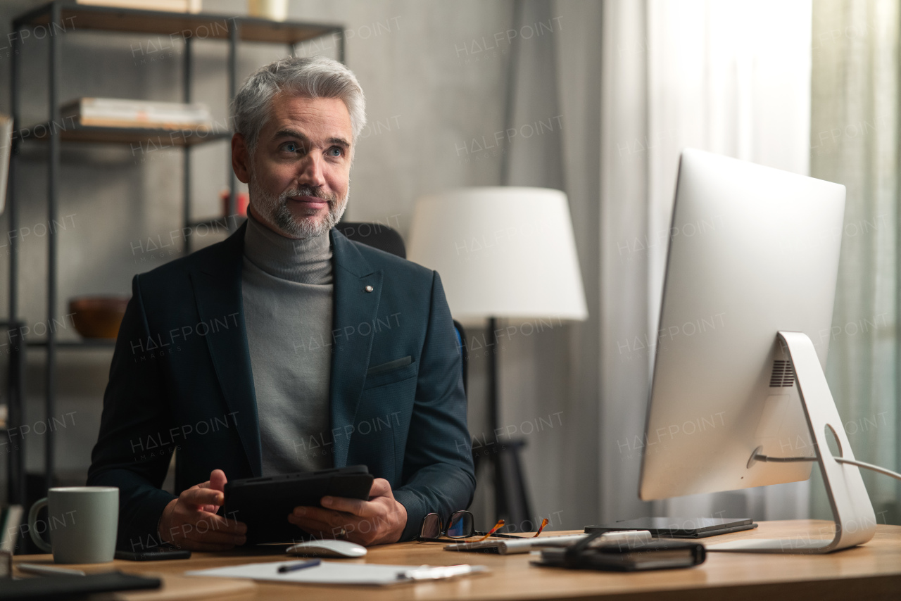 A mature businessman indoors in office, working on tablet.