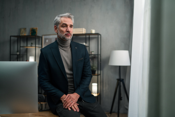 A mature sad businessman sitting on desk and thinking indoors in office.