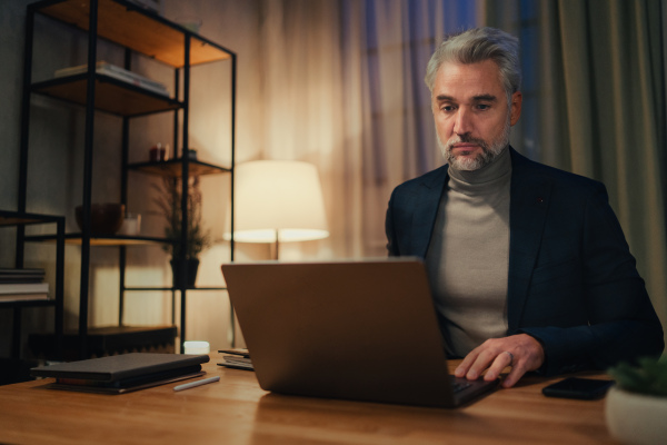 A mature businessman working on laptop at desk indoors in office at night.