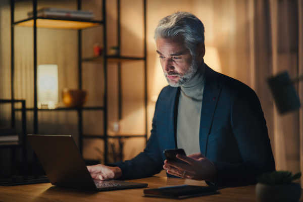 A mature businessman working on laptop at desk indoors in office at night.