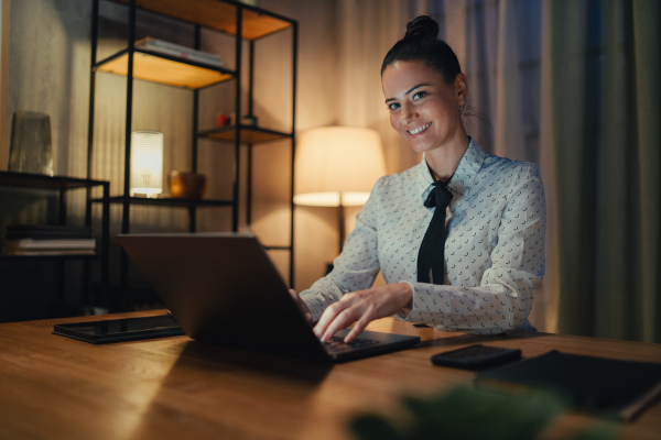 A happy mid adult business woman with diary working on laptop in office at night, looking at camera.