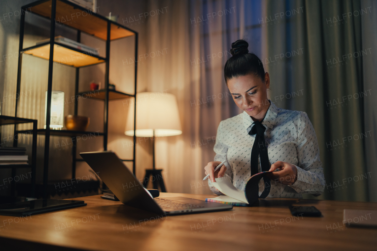 A happy mid adult business woman with diary working on laptop in office at night.