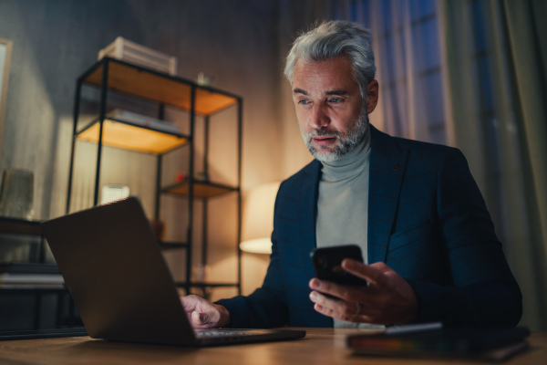 A mature businessman working on laptop at desk indoors in office at night.