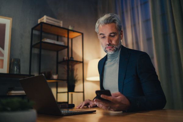 A mature businessman working on laptop at desk indoors in office at night.