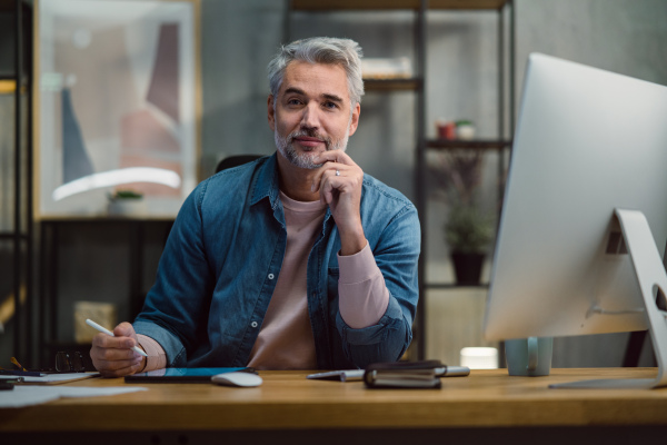 A mature man architect working on tablet at desk indoors in office, looking at camera.