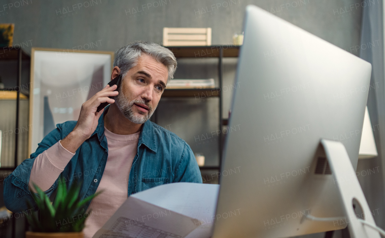 A mature man architect working on computer and making phone call at desk indoors in office.