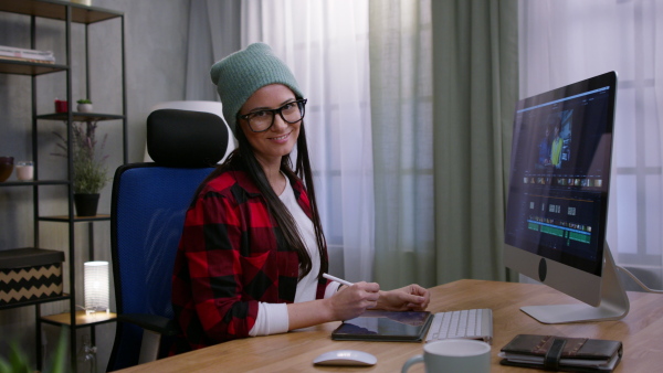 A female video editor works with footage on her personal computer indoors in creative office studio, looking at camera.