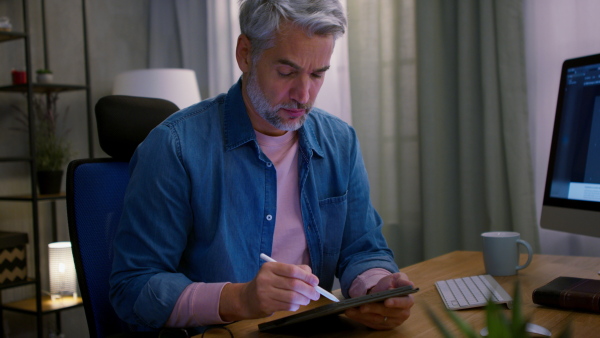 A mature businessman working on laptop at desk indoors in office, looking at camera.