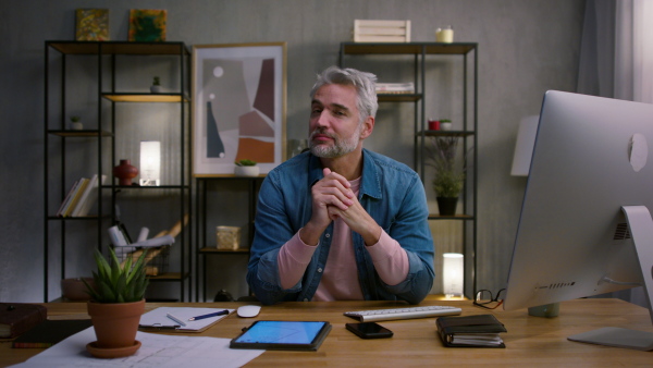 A mature businessman working on laptop at desk indoors in office, looking at camera.