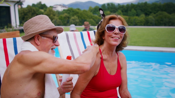 Cheerful senior couple sitting by swimming pool outdoors in backyard, using sun lotion.