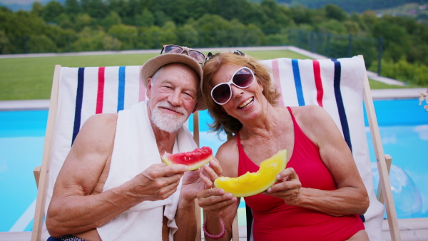 Cheerful senior couple sitting by swimming pool outdoors in backyard, looking at camera.