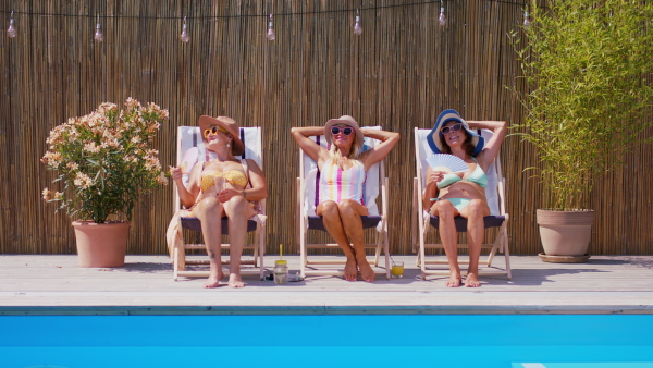 A group of cheerful senior women sitting by swimming pool outdoors.