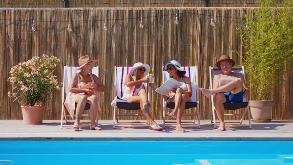 Group of cheerful seniors sitting by swimming pool outdoors in backyard, relaxing.