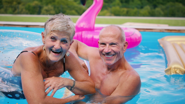 Portrait of cheerful senior couple in swimming pool outdoors in backyard, looking at camera.