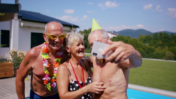 A group of cheerful seniors by swimming pool outdoors in backyard, having party and taking selfie.