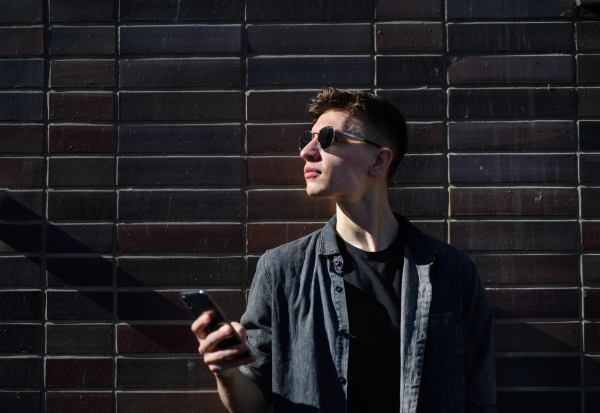 A portrait of young man standing outdoors against dark background, holding smartphone.