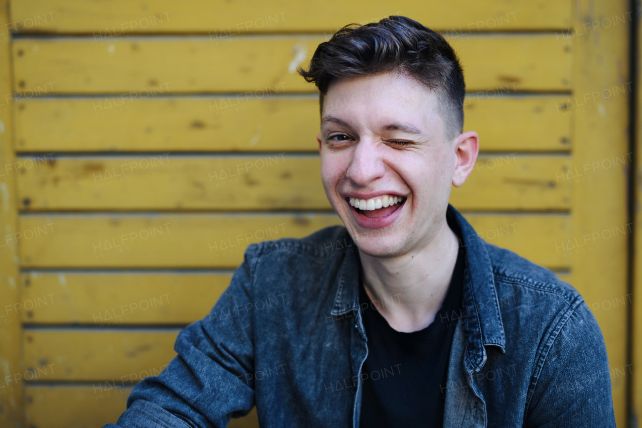 A portrait of young man standing outdoors against yellow background, winking.