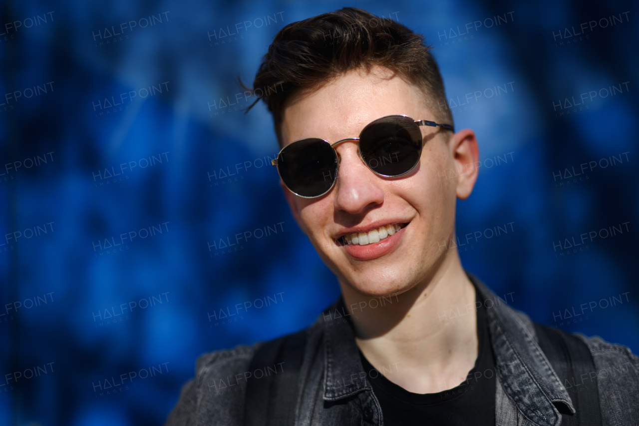 A portrait of young man with sunglasses standing outdoors against blue background, looking at camera.