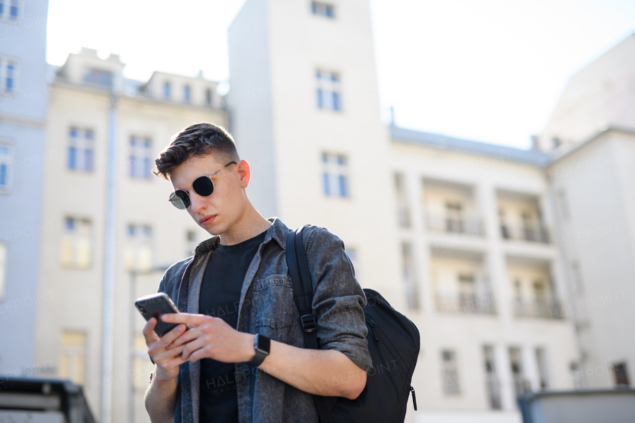 A portrait of young man standing outdoors in city, using smartphone.