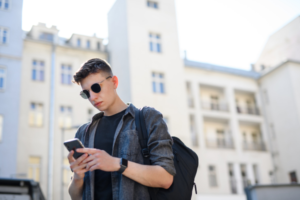 A portrait of young man standing outdoors in city, using smartphone.