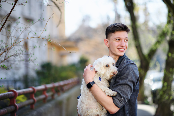 A portrait of young man walking outdoors on street in city, holding dog.