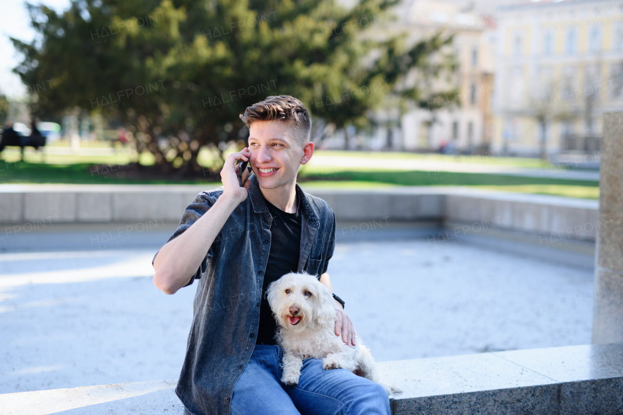 A portrait of young man with dog outdoors in city, using smartphone.