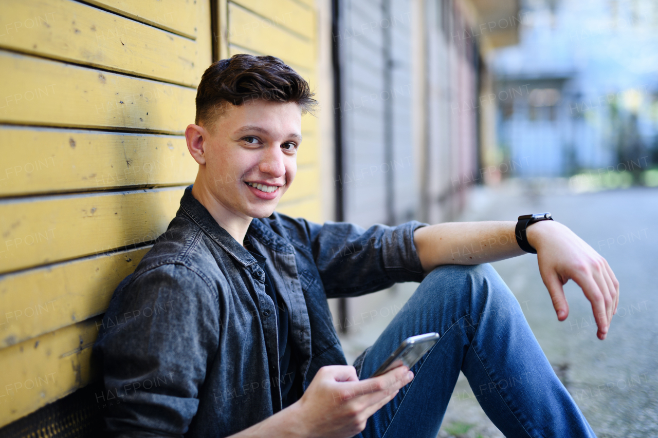 A portrait of young man with smartphone sitting outdoors in city street, looking at camera.