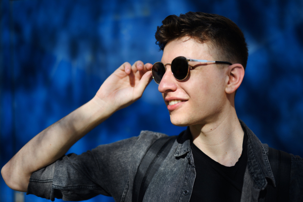 A portrait of young man with sunglasses standing outdoors against blue background.