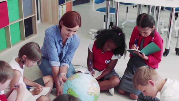 A group of small school kids with teacher sitting on the floor in class, learning geography.